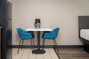 Ein kleines Zimmer mit einem weißen Tisch, zwei blauen Stühlen, einer Kaffeemaschine, einem Bett und einem Kühlschrank.