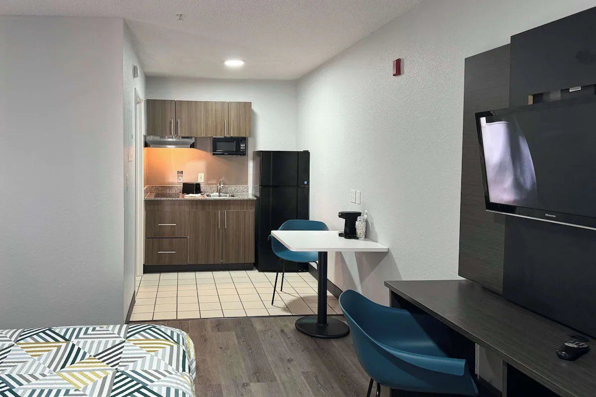 Een kleine hotelkamer met een kitchenette, een bed met beddengoed met een patroon, een televisie en een eethoekje.