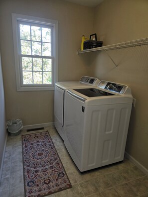 A laundry room with a front-loading washer and dryer, a window, a shelf with bottles, and a patterned rug.