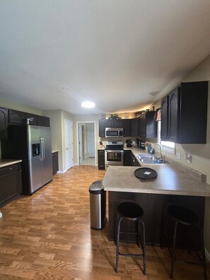 A kitchen with a stainless steel refrigerator, a microwave, an oven, and a sink. There is a trash can and a bar stool.