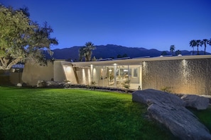 A modern house with a well-maintained lawn, large rocks, and a mountain backdrop. - The Cascades Retreat: 3 Bedroom Home with Private Pool and Mountain Views (Palm Springs)
