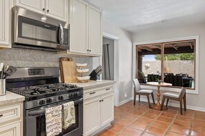 A kitchen with a stove, microwave, and cabinets, a dining area with chairs and a table, and a view through a window. - Vista El Quetzal - Central Palm Springs (Palm Springs)