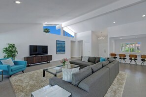 A modern living room with a sectional sofa, a flat-screen TV, and a large window providing natural light. - Twin Palms Tranquility (Palm Springs)