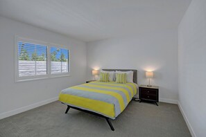 A bedroom with a bed, two bedside lamps, a window with blinds, and a carpeted floor. - Twin Palms Tranquility (Palm Springs)