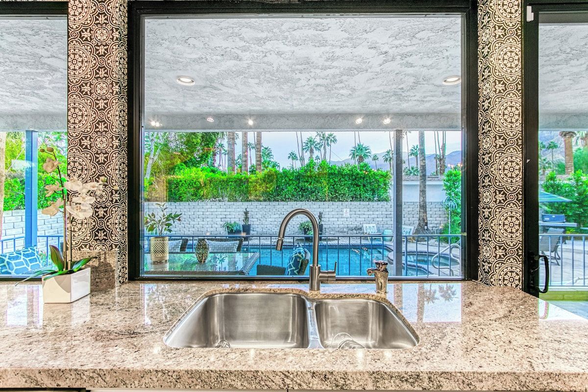 A kitchen with a large window overlooking a swimming pool and greenery.