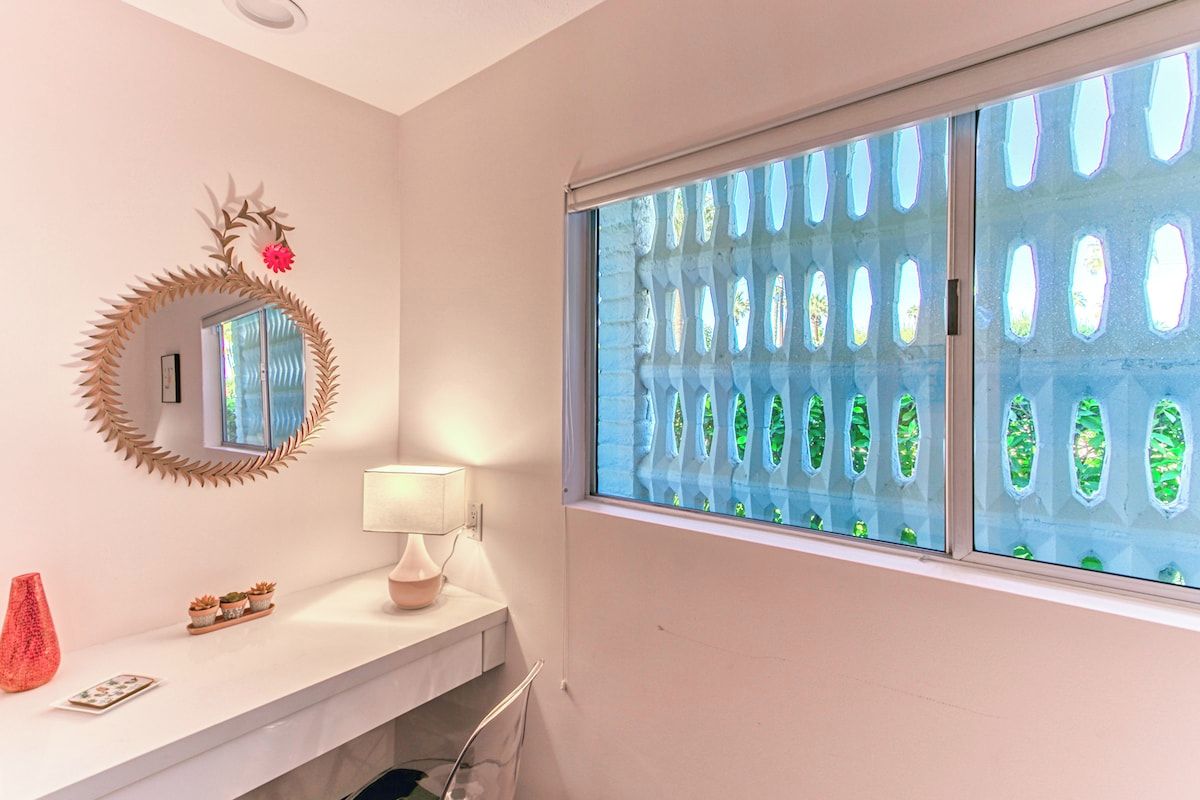 Casa de banho com toucador branco, espelho com decoração de folhas, janela com vista para folhagem verde e um vaso vermelho.