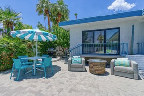 An outdoor patio with a table and chairs, a stone fire pit, and a blue and white umbrella. - Luxury Deepwell Stunner (Palm Springs)