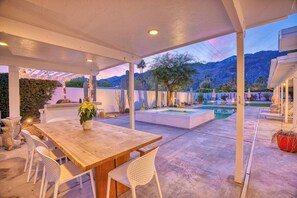 An outdoor patio with a wooden table and chairs, a pool, and a mountain backdrop. - Puerta Del Sol - Tahquitz River Estates (Palm Springs)