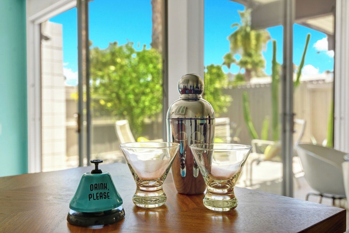 A table with a cocktail shaker, two shot glasses, and a 'Drink, please' sign.