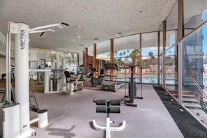 Ein Fitnessraum mit unterschiedlichen Trainingsgeräten, einem großen Fenster mit Blick auf ein Schwimmbecken und eine Palmenlandschaft.