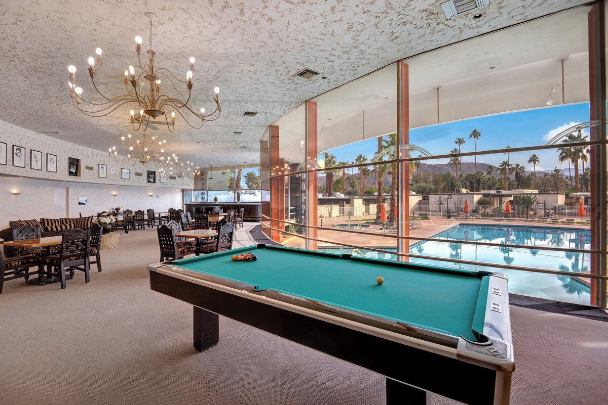 A pool table in a room with large windows overlooking a pool and palm trees.