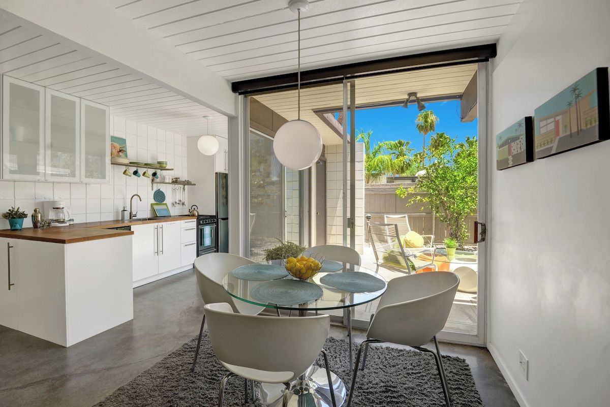Una cocina moderna con muebles blancos, una mesa de comedor de vidrio y vistas al exterior.