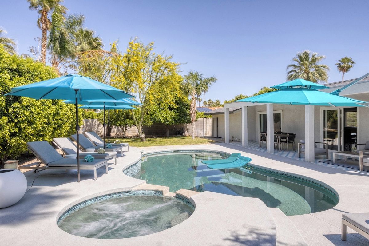 A swimming pool area with lounge chairs and umbrellas, surrounded by greenery and a building with large windows.