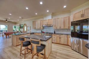A spacious kitchen with wooden cabinets, a central island with bar stools, a stainless steel refrigerator, and a pool table in the background. - PGA West Paradise by Oranj Palm (La Quinta)
