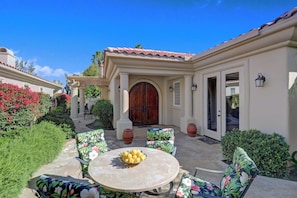 A patio with outdoor furniture and a table, surrounded by greenery and a building with a tiled roof. - PGA West Paradise by Oranj Palm (La Quinta)