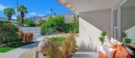 Un patio moderno al aire libre con suelo de hormigón, una zona de estar con mesa de madera y sillas naranjas, y vistas a montañas y palmeras.