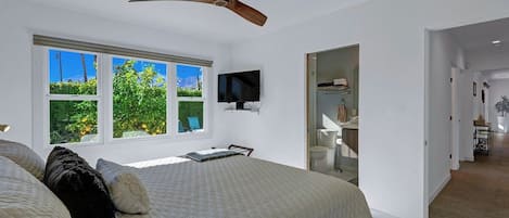 A bedroom with a bed, a ceiling fan, a TV, and a window with a view of greenery.