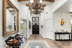 A hallway with a large ornate mirror, a chandelier, a black door, and a console table with decorative items. - Casa Sueños - Andreas Hills (Palm Springs)