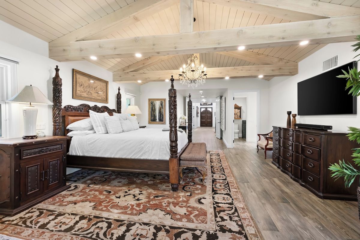 A bedroom with a large bed, wooden headboard, a chandelier, a dresser, and a television.