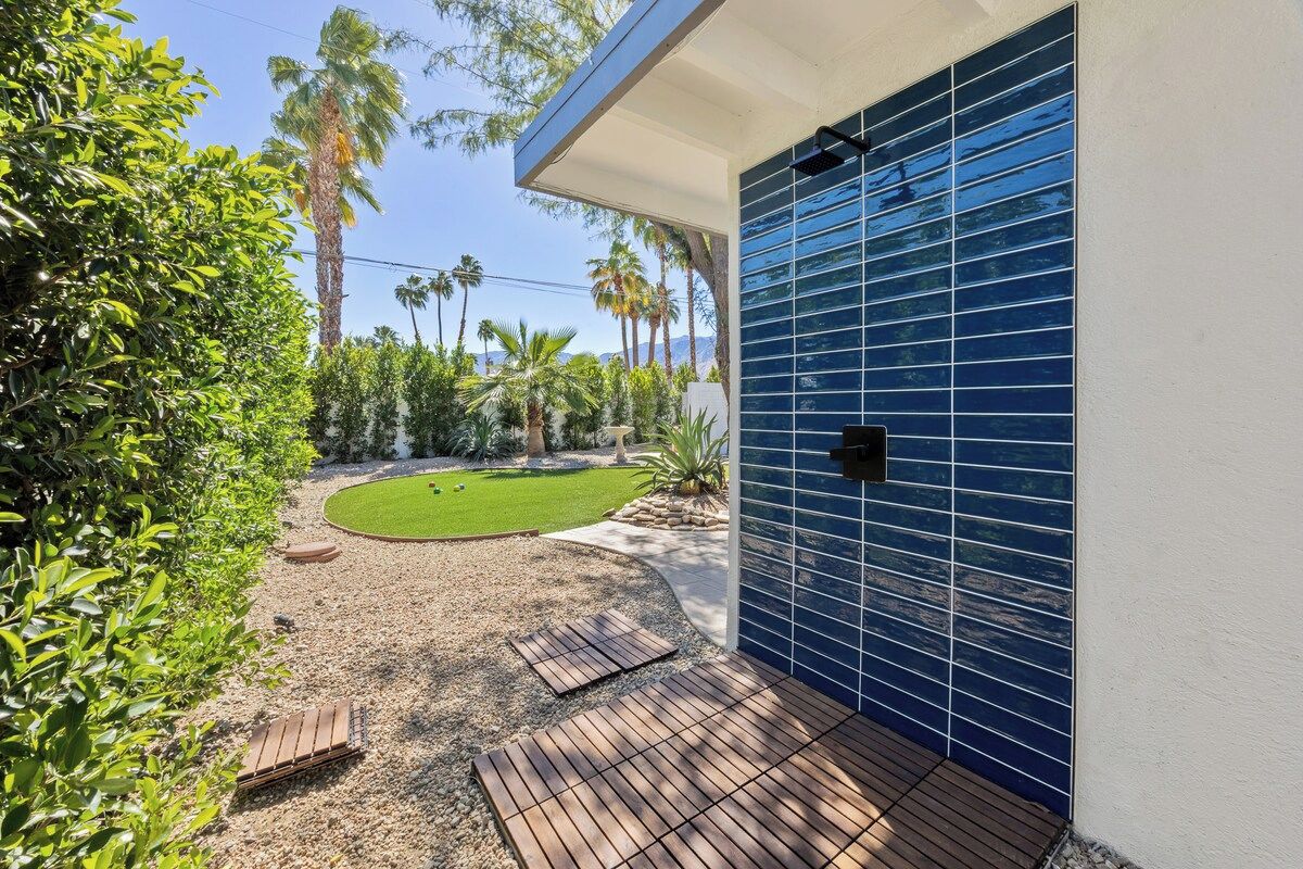 Une terrasse extérieure moderne, dotée d’une plateforme en bois, d’un mur recouvert de carreaux bleus et d’une pelouse bien entretenue agrémentée de palmiers.