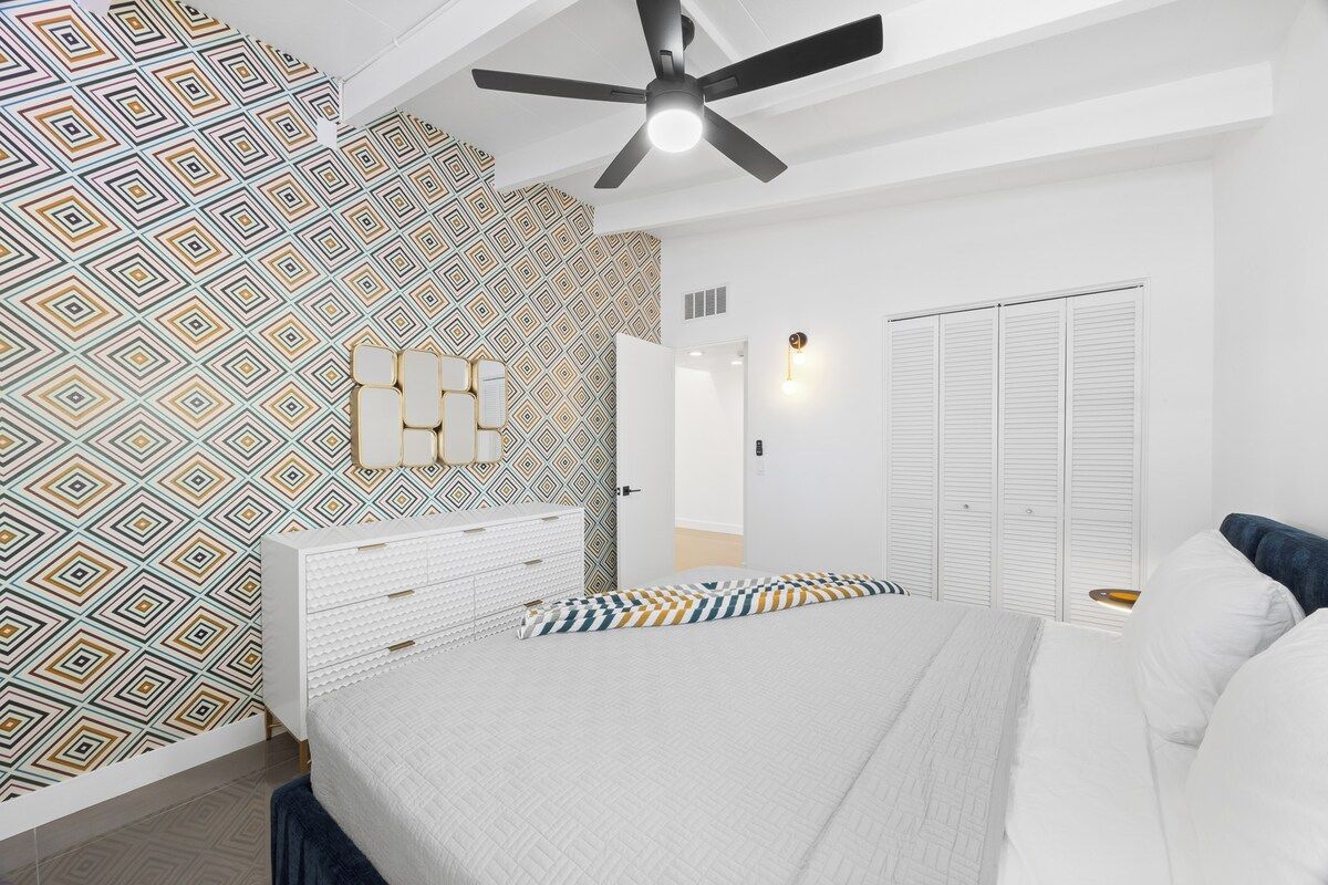 A bedroom with a patterned wall, a bed with white bedding, a white dresser, and a ceiling fan.