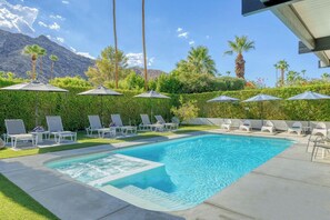 Ein Swimmingpool mit Liegestühlen und Sonnenschirmen, umgeben von Grünflächen mit Blick auf Berge.
