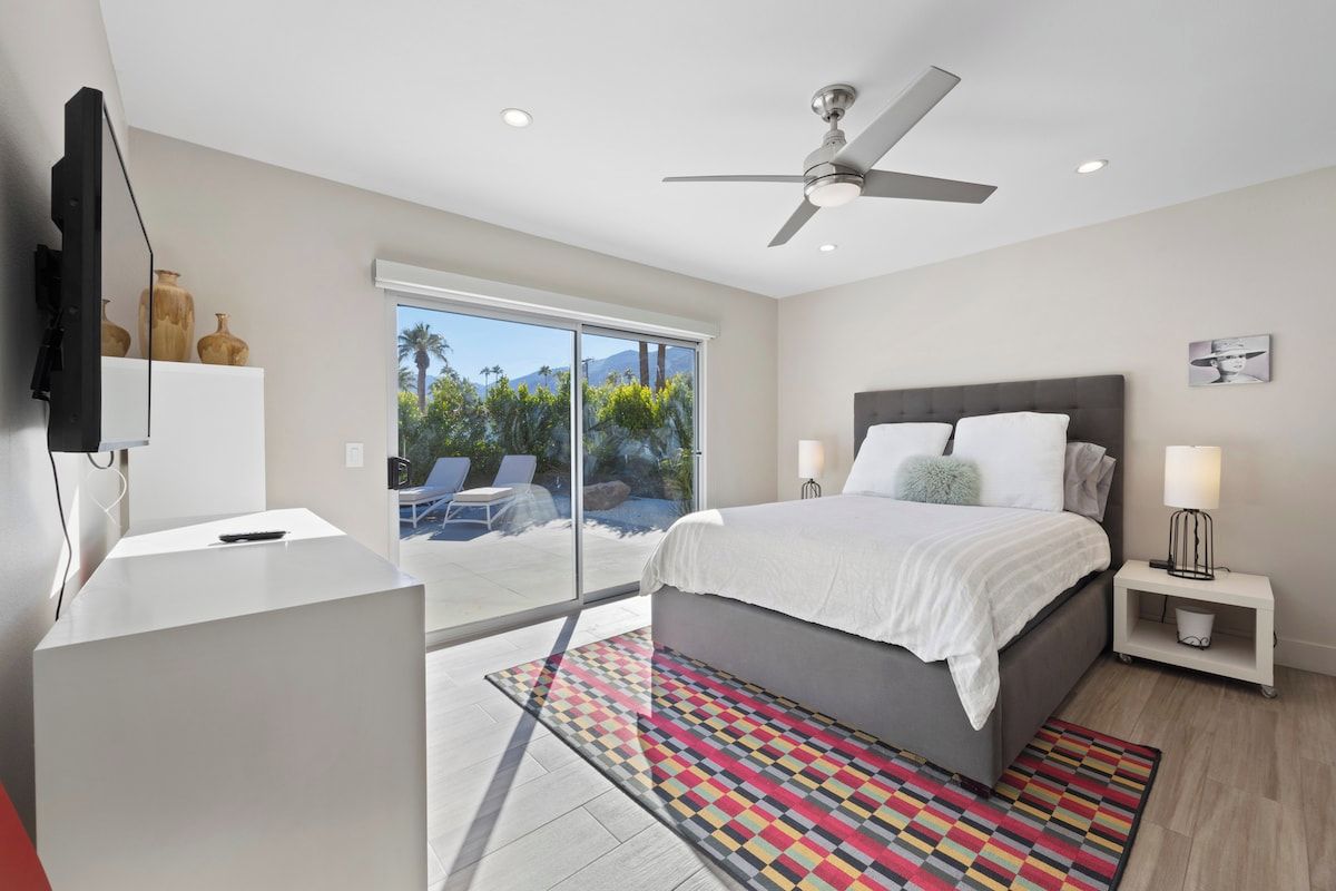 A bedroom with a bed, a ceiling fan, a TV, a desk, and a view of the outdoors.