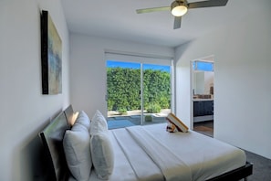 Ein modernes Schlafzimmer mit einem großen Bett, einem Deckenventilator und Blick auf einen üppigen Garten durch eine Schiebeglasdoor.