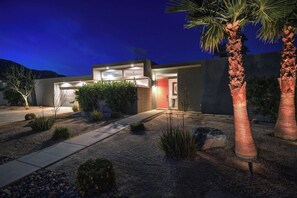 A modern house with a garage, a well-lit entrance, and a landscaped garden with palm trees and shrubs. - Oranj Door Modern - Warm Sands (Palm Springs)