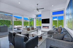 A modern living room with a fireplace, large windows, and a ceiling fan. - Oranj Door Modern - Warm Sands (Palm Springs)