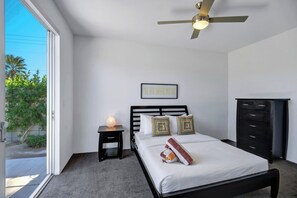 A bedroom with a bed, a nightstand, a dresser, a ceiling fan, and a window with a view of greenery. - Oranj Door Modern - Warm Sands (Palm Springs)