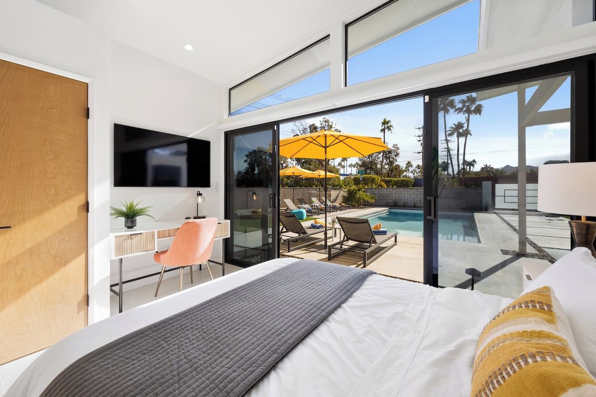 A modern hotel room with a large bed, a TV, a desk with a chair, and a view of a swimming pool and palm trees.