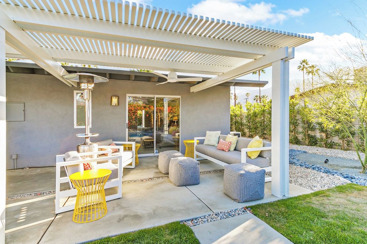 A covered outdoor seating area with a sofa, chairs, and ottomans, a pergola, and a view of a pool and garden.