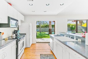 A modern kitchen with white cabinetry, a built-in oven, and a large window overlooking a backyard. - Dreamville Palm Springs by Oranj Palm (Palm Springs)