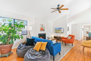 A modern living room with a fireplace, a ceiling fan, and a large window with a view of palm trees. - Dreamville Palm Springs by Oranj Palm (Palm Springs)