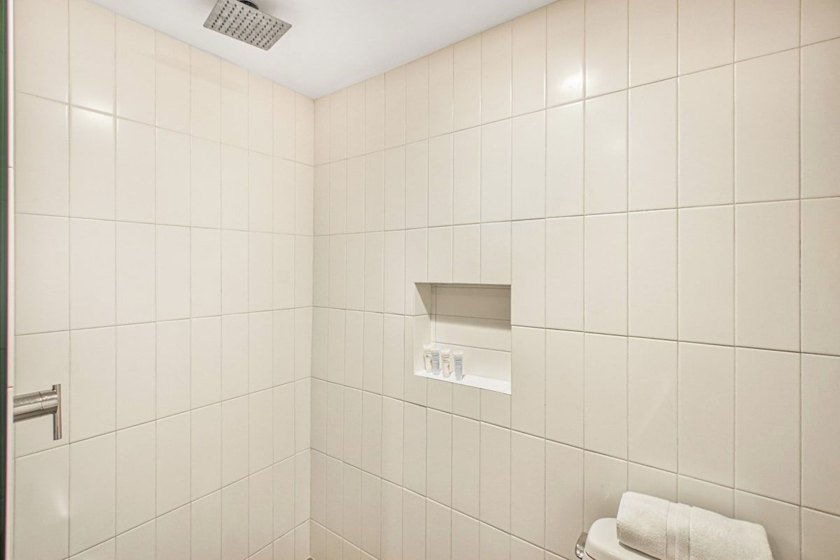 Ein Badezimmer mit weißen Fliesen, einer Duschbrause, einem Handtuchhalter und einem Ablagebereich für Toilettenartikel.