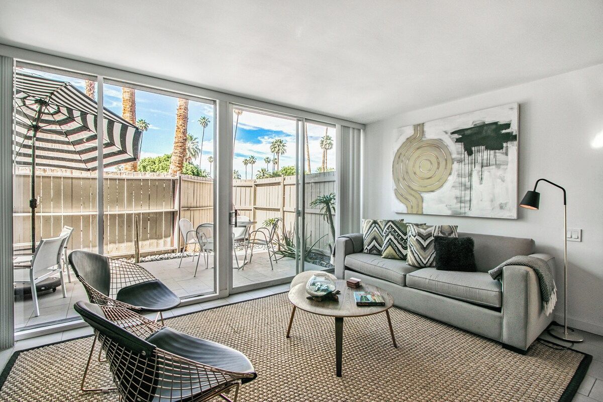 A modern living room with a sofa, chairs, a coffee table, and a large window with a view of palm trees.