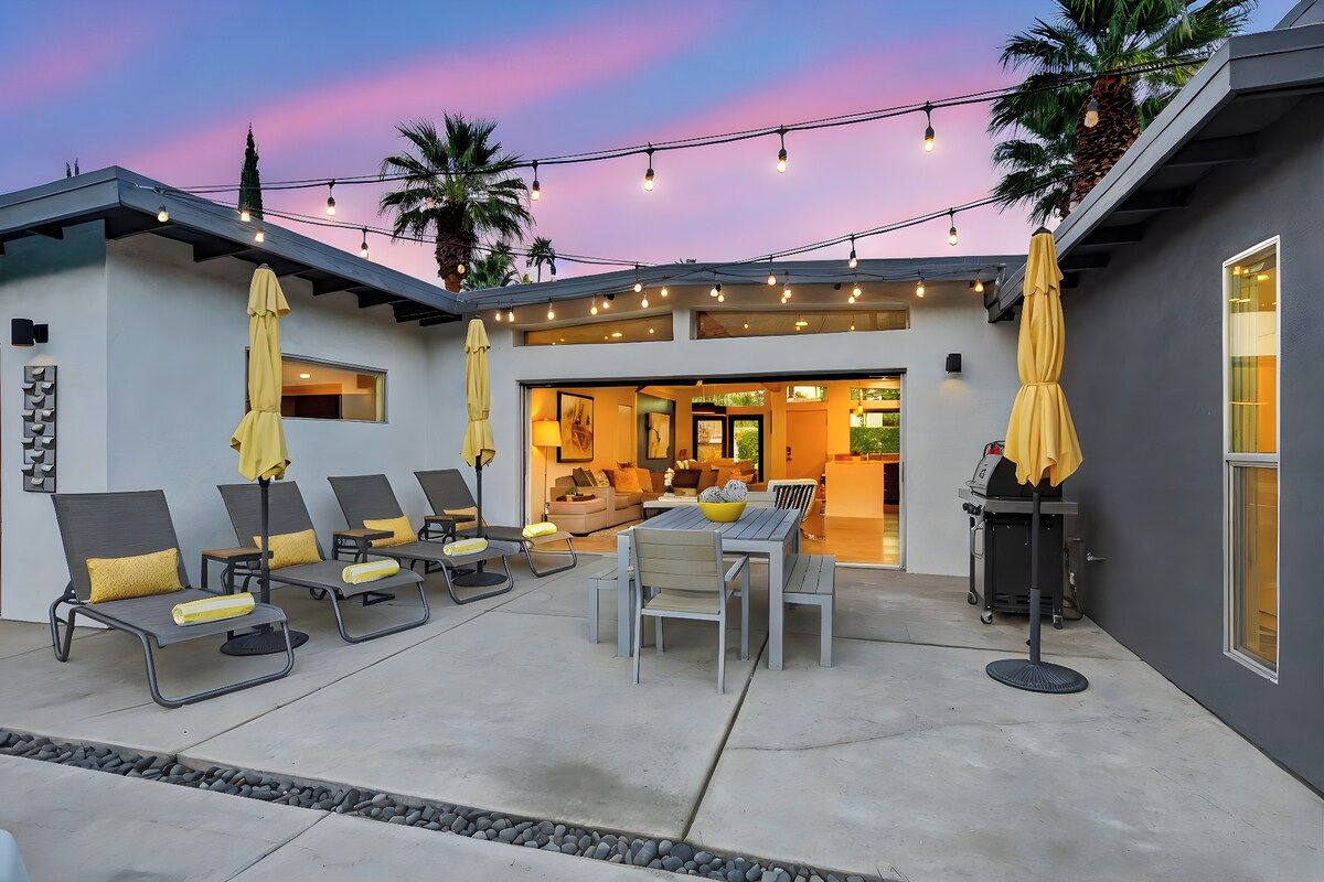 An outdoor patio with lounge chairs and a dining table, string lights overhead, and a view into the house.