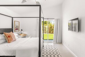 A modern bedroom with a four-poster bed, a nightstand with a lamp, a framed picture, and a view of a green lawn through a sliding glass door. - Parkview Bungalow - South PS (Palm Springs)