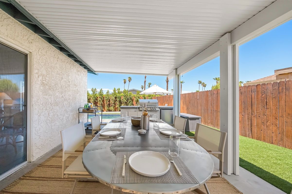 An outdoor patio with a round table set for dining, a grill, and a view of a wooden fence and greenery.