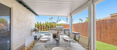 An outdoor patio with a round table set for dining, a grill, and a view of a wooden fence and greenery.