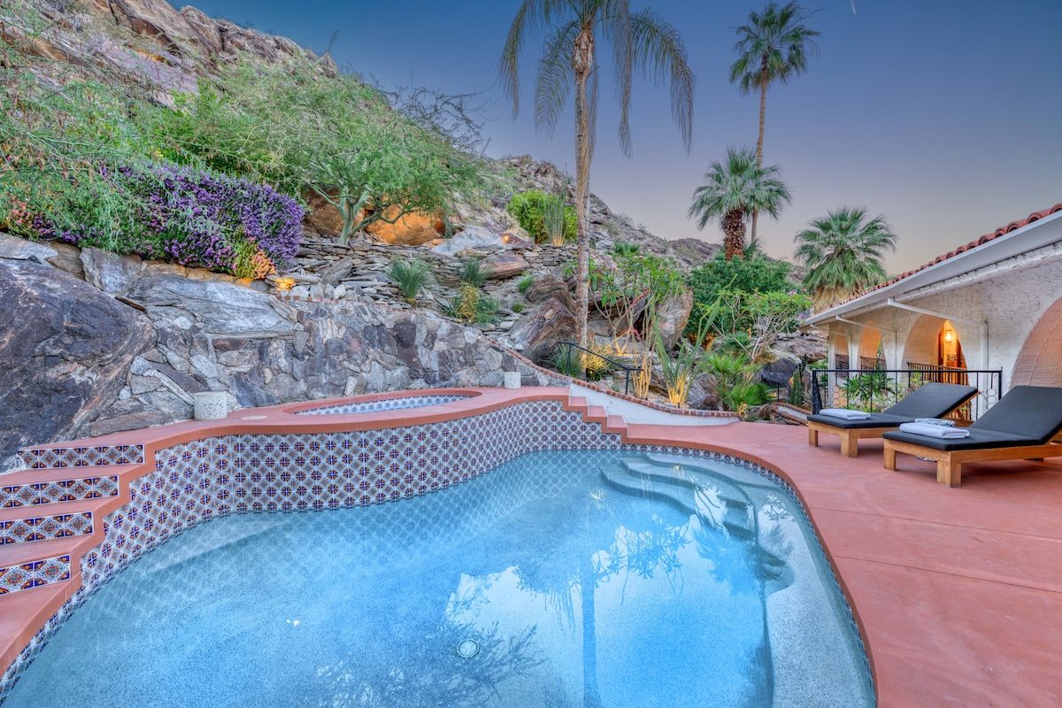 A swimming pool with a tiled edge, surrounded by rocks and plants, with lounge chairs and a building in the background.