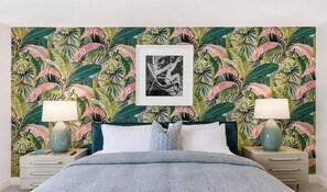Ein Schlafzimmer mit einem Bett, zwei Nachttischen, einem Beistelltisch, einem gerahmten Bild und Tapete mit tropischem Blattmuster.