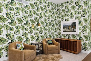 A room with a tropical leaf wallpaper, two armchairs, a small round table, a cabinet, and a framed picture on the wall. - Villa Palmeras (Palm Springs)