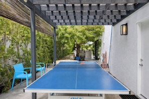 An outdoor ping pong table with blue netting, surrounded by blue chairs, under a pergola with a woven roof. - Villa Palmeras (Palm Springs)