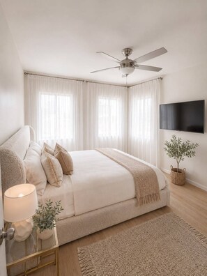 Ein Schlafzimmer mit einem Bett, einem Deckenventilator, einem Fernseher und einem Fenster mit Vorhängen.