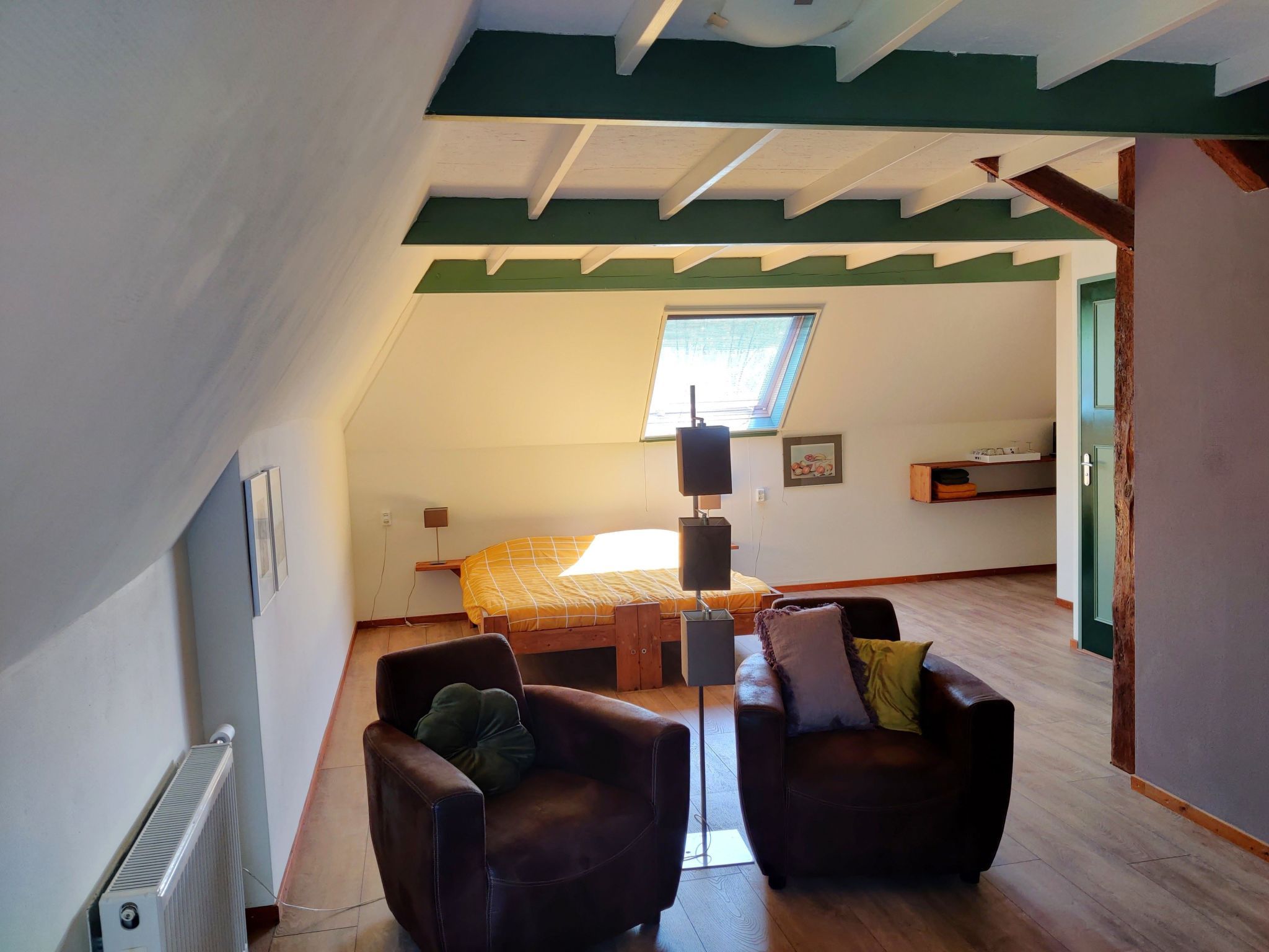 A room with a sloped ceiling, a bed with a yellow bedspread, two leather chairs, a radiator, and a window with blinds.