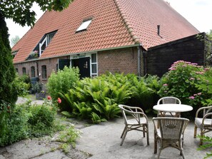 Een gebouw van baksteen met een dak van rode tegels, een tuin met varens en roze bloemen, en rieten meubels.