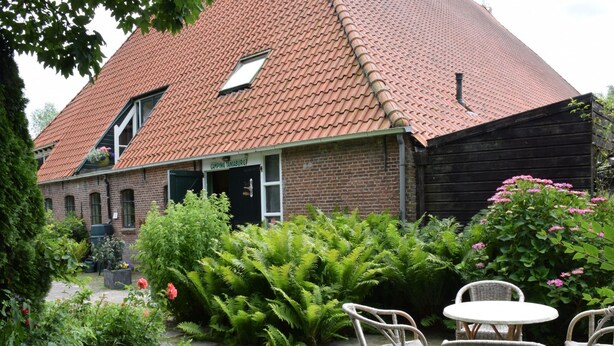 Ein Backsteinhaus mit rotem Ziegeldach, ein Garten mit Farne und rosa Blüten sowie Korbmöbel.