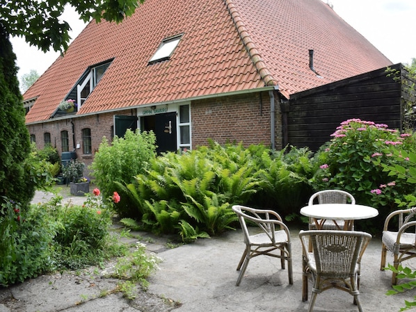 A brick building with a red-tiled roof, a garden with ferns and pink flowers, and wicker furniture. - Taniaburg Gästezimmer 2 by Interhome (Leeuwarden)